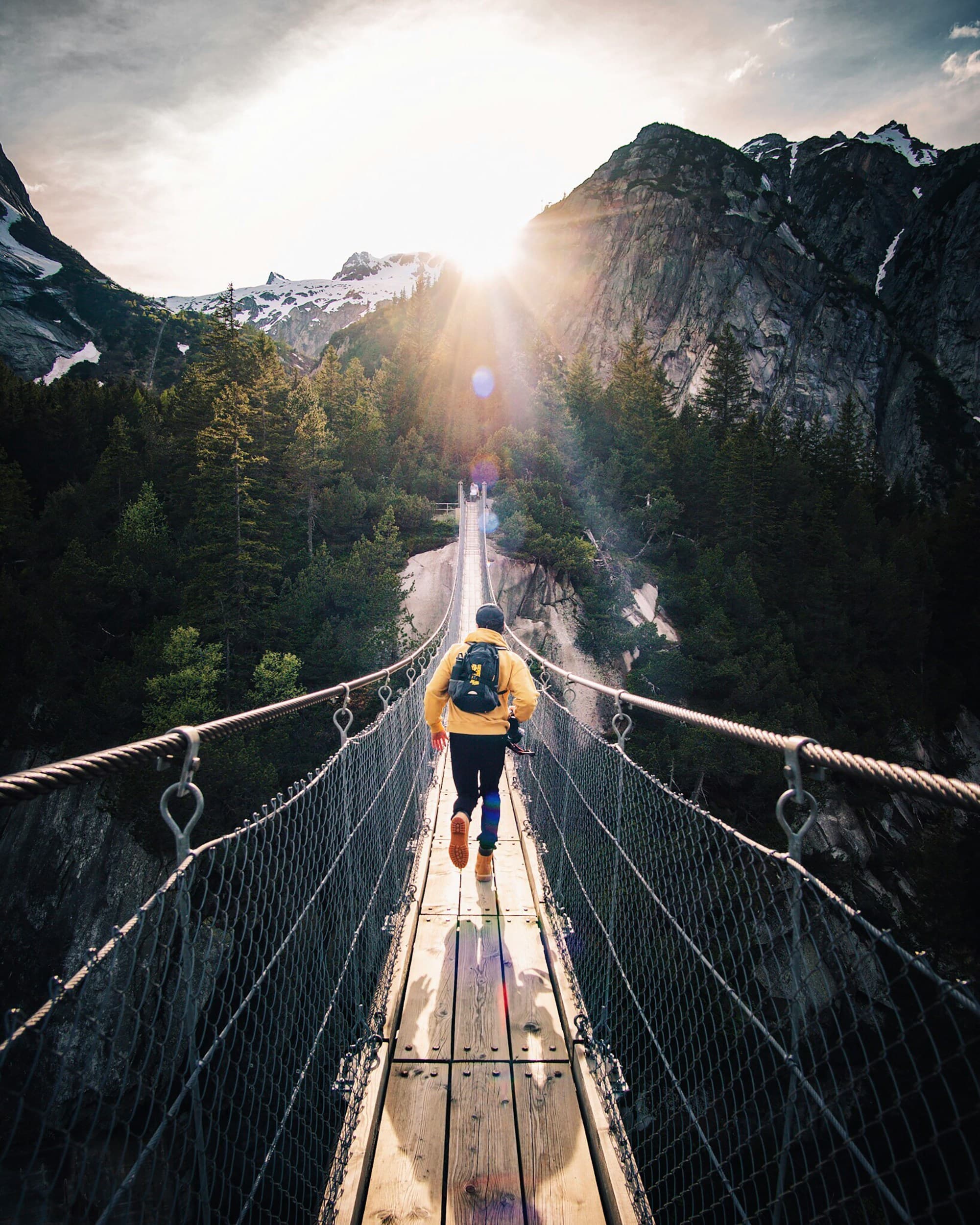 A hiker crossing a suspension bridge toward sunlit mountain peaks